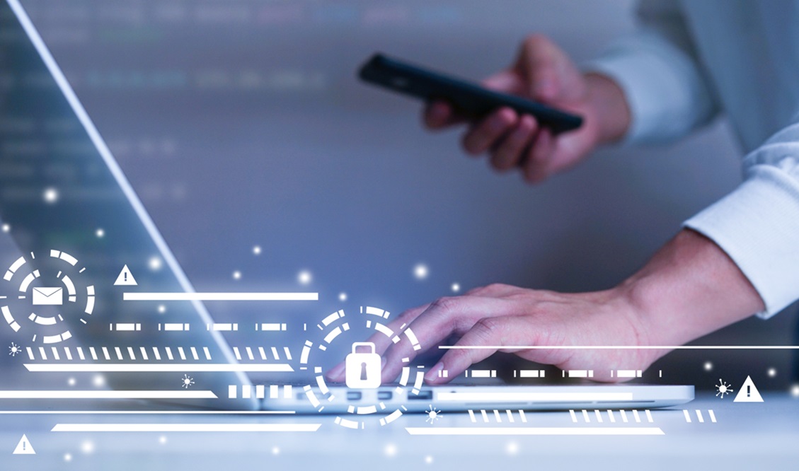 A man typing with one hand on a laptop working on a cyber security risk register, and a mobile phone in his other hand 